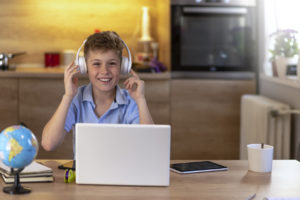 Photo of smiling student sitting at the kitchen table, online learning with laptop and smartphone during the day at home, looking at camera with smile. Teenage boy with headphones using laptop at table in room. 11 years old boy sitting behind a laptop doing online school while smiling.