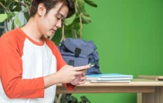A young student at home who is distracted by his phone while doing online school.